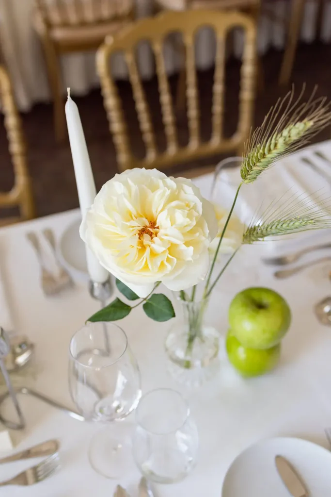 Close-up of a non-traditional wedding table setting featuring a single blooming white garden rose David Austin, fresh green apples, and delicate wheat stems. Tailor-made floral design at Palácio do Freixo by Mande uma Flor.