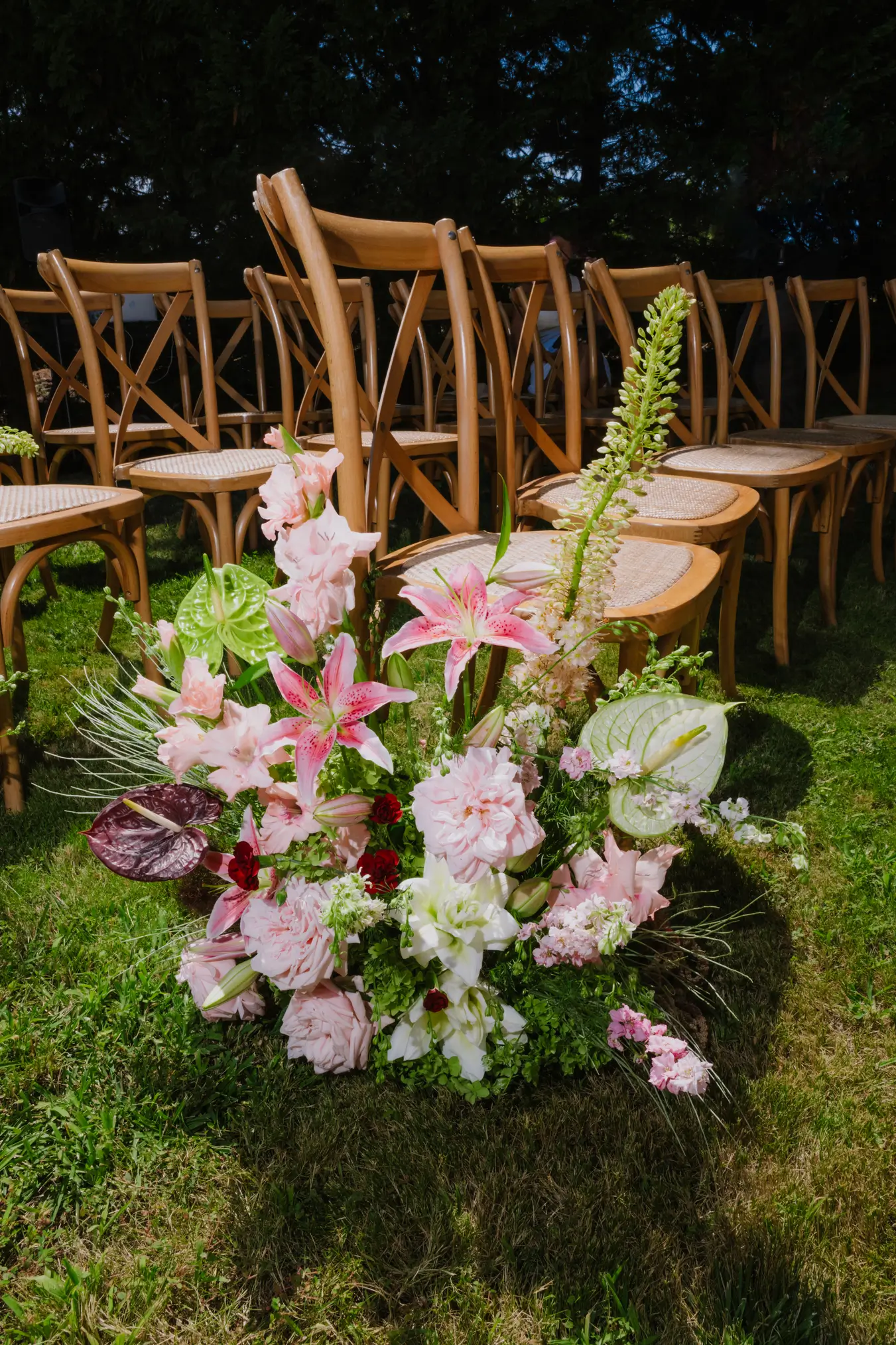 Home 7 Lush and sculptural ground floral arrangement featuring pink lilies, anthuriums, and roses, set next to wooden cross-back chairs for an outdoor wedding ceremony. Tailor-made design by Mande uma Flor.
