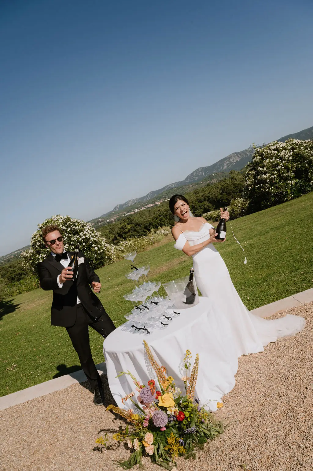 Home 3 A joyful couple popping champagne by a glass tower outdoors, accented with a vibrant, tailor-made ground floral arrangement. Destination wedding in Portugal featuring bespoke styling by Mande uma Flor.