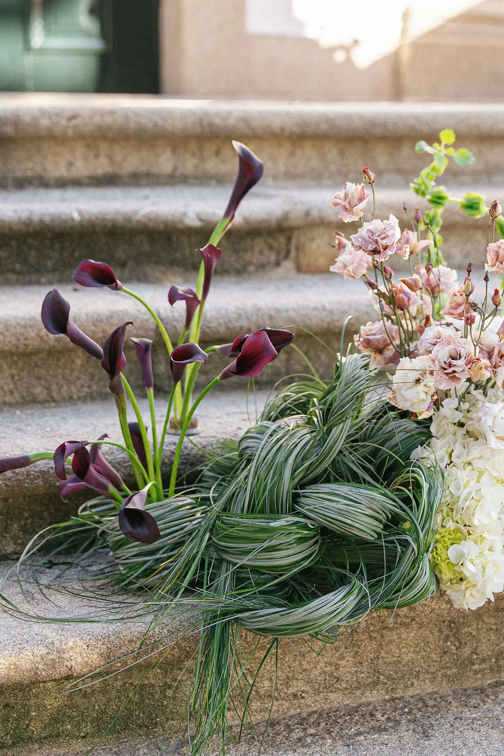 Home 5 Artistic and non-traditional floral arrangement decorating stone steps, featuring sculptural dark purple calla lilies, woven green grass, and soft pink blooms. Tailor-made wedding styling.